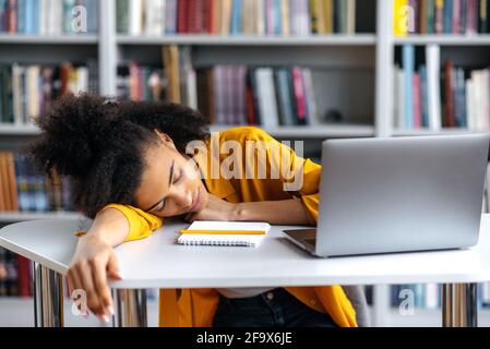 Una studentessa afro-americana dormiva stanca addormentandosi ad un tavolo durante una conferenza ad un college. La ragazza riposa dallo studio, chiudendo gli occhi e rilassandosi alla sua scrivania Foto Stock