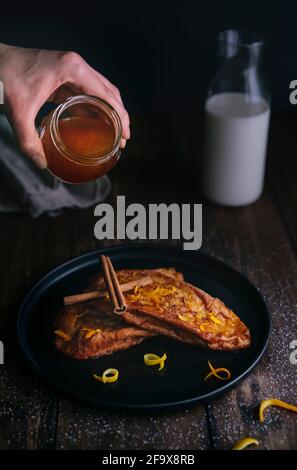 Versate a mano il miele da un vaso di vetro sul pane fritto fette con due bastoncini di cannella e bucce di arancia e limone in un piatto nero su una tavola di legno scuro Foto Stock