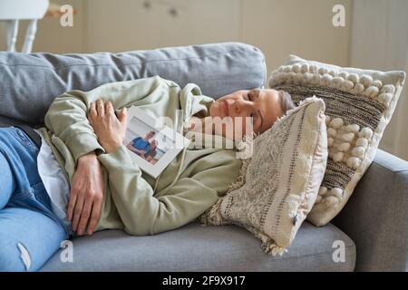 Triste donna di mezza età con foto di famiglia con i suoi figli in cornice mantenendo gli occhi chiusi e sognando mentre si sdraia sul divano a casa. Focalizzazione selettiva sulle donne anziane Foto Stock