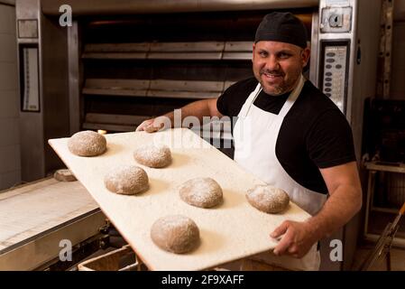 panettiere in panetteria sorridente e in posa con mensola con pane crudo impastato prima della cottura. concetto di preparazione manuale tradizionale del pane Foto Stock