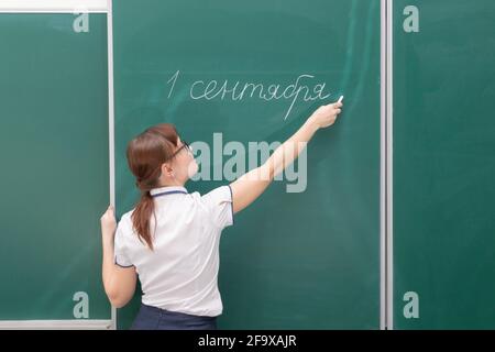 Insegnante di scuola giovane donna in una blusa bianca in classe su una lavagna verde scrive con gesso. Traduzione - 1 settembre. Ritratto Foto Stock