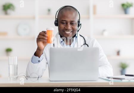 Medico che raccomanda la medicina. Terapeuta afroamericana in cuffie che mostrano il vaso di pillole per lo schermo del computer portatile Foto Stock