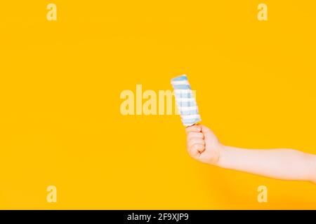 Mano dei bambini che tiene un gelato a strisce su un bastone davanti a uno sfondo giallo. Foto Stock