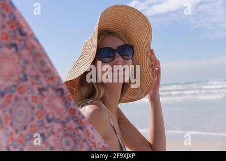 Donna caucasica sorridente che indossa bikini e cappello seduto sul ponte sedia che guarda la macchina fotografica sulla spiaggia Foto Stock