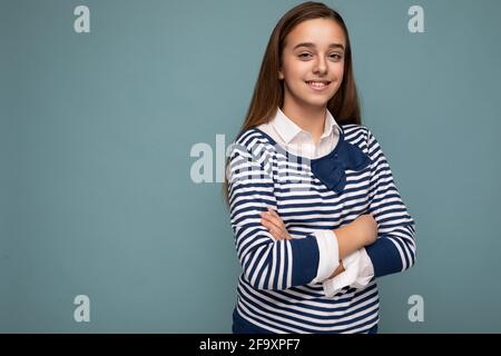 Scatto di bella bruna positiva sorridente bambina indossando a strisce maniche lunghe isolate sulla parete blu di fondo che attraversa le mani e. Foto Stock