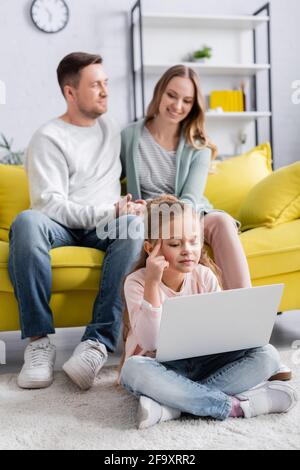 Ragazza che usa il laptop durante la formazione online vicino ai genitori su offuscato sfondo Foto Stock