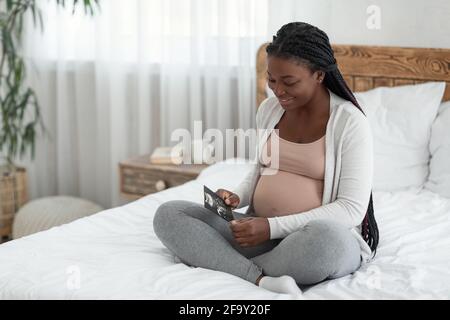 Donna incinta africana felice seduta sul letto con ultrasuoni bambino Acquisisci immagine Foto Stock
