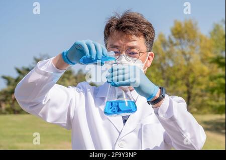 Ritratto di uno scienziato asiatico di metà anno che indossa un abito da laboratorio bianco e guanti e tiene in mano un contenitore sperimentale. Foto Stock