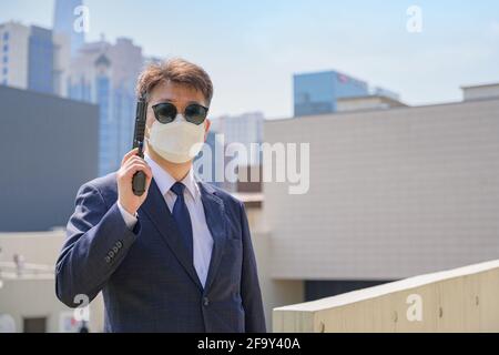 Un maschio asiatico che tiene una pistola indossando occhiali da sole e una maschera sullo sfondo della città. Foto Stock