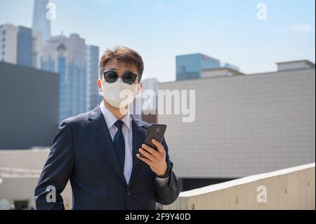 Uomo d'affari asiatico che tiene smartphone indossando occhiali da sole e maschera in città. Foto Stock