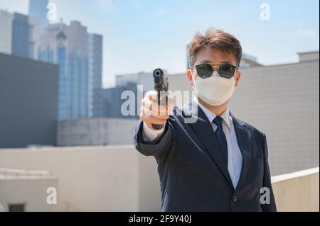 Un maschio asiatico che tiene una pistola indossando occhiali da sole e una maschera sullo sfondo della città. Foto Stock