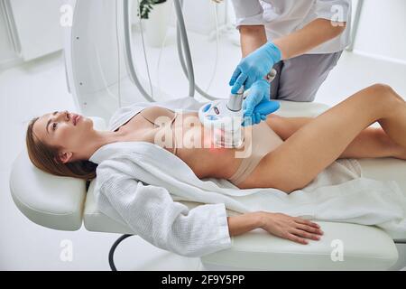 Affascinante adorabile donna sdraiata sul tavolo della medicina mentre si ottiene cavitazione trattamento di contornamento del corpo in medicina clinica di bellezza Foto Stock