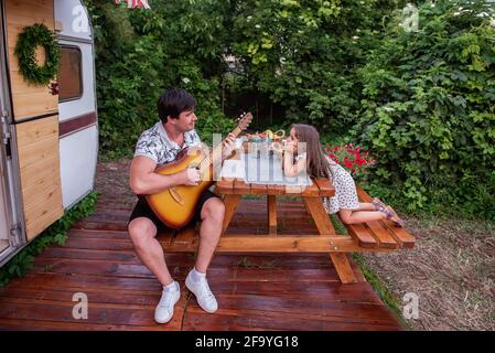 Giovane bruna padre sta suonando canzoni chitarra per piccola bella figlia. L'uomo e la ragazza sono seduti al tavolo di legno nel cortile godendo il mus Foto Stock