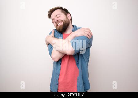 Affascinante uomo bearded si abbraccia, chiude gli occhi e i sogni d'amore, sorridendo dolcemente. Sfondo bianco. Foto Stock