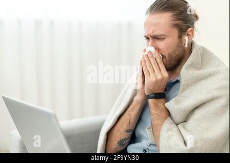Il ragazzo ha un raffreddore e un'influenza, si starnutisce in carta tissue seduto a casa coperto di coperta. Un giovane ha una malattia virale preoccupata per i suoi malati, seduto sul divano con un laptop Foto Stock