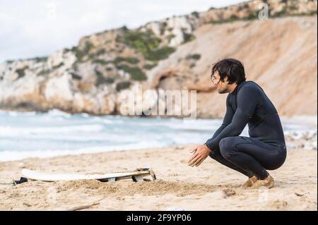 Vista laterale di un uomo bagnato accovacciato che indossa un costume da immersione. Maschio sulla spiaggia con la sua tavola da surf riposante e guardando il mare dopo il surf. Foto Stock