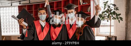 studenti multietnici in maschere mediche, abiti di laurea e cappelli con diploma, striscione Foto Stock