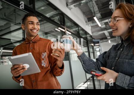 Studente afroamericano che tiene una tavoletta digitale e tazze di carta graffetta con felice compagno di classe Foto Stock