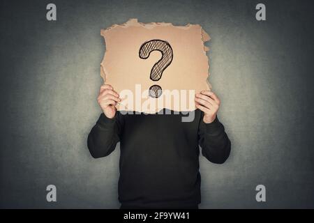 L'uomo anonimo copre la testa utilizzando un foglio di cartone strappato, come una maschera, con un punto interrogativo disegnato. Persona incognito nascosta, isolata sulla parete grigia posteriore Foto Stock