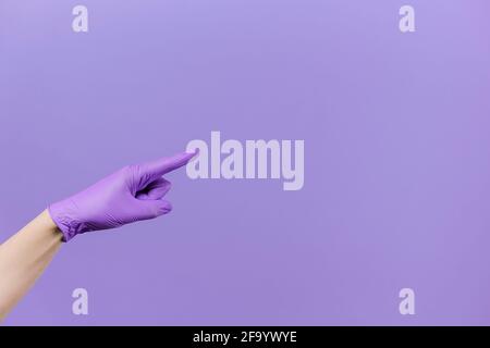 Il dito maschio che punta verso lo spazio di copia, indossa guanti in lattice di protezione medica, isolati sulla parete di fondo viola dello studio con spazio di copia Foto Stock