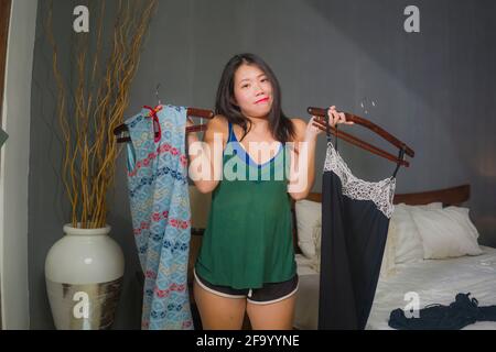 Ritratto naturale di giovane felice e dolce donna coreana asiatica provando i vestiti differenti che osservano nello specchio della camera da letto che decide che cosa nuovo vestito per datare Foto Stock