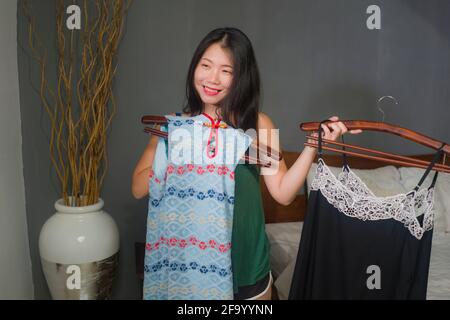 Ritratto naturale di giovane felice e dolce donna giapponese asiatica provando i vestiti differenti che osservano nello specchio della camera da letto che decide che cosa nuovo vestito per datare Foto Stock