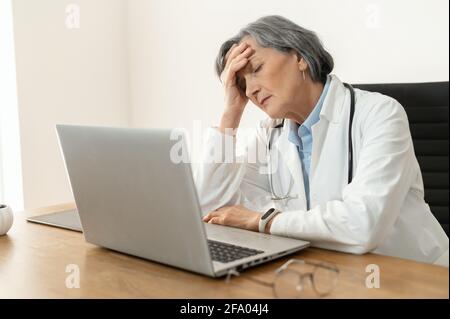 Stanco ha stressato la dottoressa anziana in un cappotto di laboratorio seduto in ufficio di fronte a un computer portatile, sentendosi scurnout al lavoro, irrequieto dopo non avere giorni fuori o esaurito, e addormentarsi Foto Stock