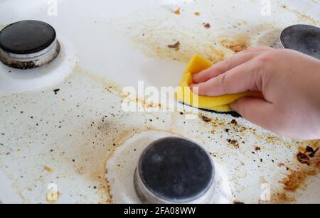 Una mano con una spugna di lavaggio gialla lava la superficie grassa molto sporca della stufa a gas. Dopo la spugna, rimane una traccia pulita. Foto Stock