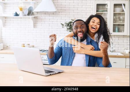 Una coppia multirazziale molto contentissima in cucina a casa celebrando la vittoria, si siede in abbracci davanti al laptop, guarda la telecamera e grida felicemente, sorpresi uomo afroamericano e donna Foto Stock