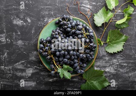 Uve nere in piastra con pianta di vite su tavolo scuro vista dall'alto. Cibo di uva succosa dolce su fondo nero di cemento piatto posato. Foto Stock
