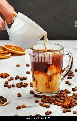 Una mano dell'uomo versa la panna in una tazza trasparente con caffè sullo sfondo di chicchi di caffè sparsi, fette di limone essiccate e stelle di anice. Foto Stock