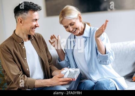 Felice coppia di mezza età innamorata. Un marito gioioso e amorevole dai capelli grigi dà una bella scatola con un regalo costoso per il loro anniversario di matrimonio o il suo compleanno, moglie affascinante è felice e sorridente Foto Stock