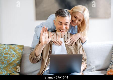 Comunicazione di videochiamata. Felice coppia sposata caucasica senior seduta sul divano in abiti casual, utilizzando il laptop per chattare online con amici o familiari tramite videoconferenza, saluto, sorridente Foto Stock