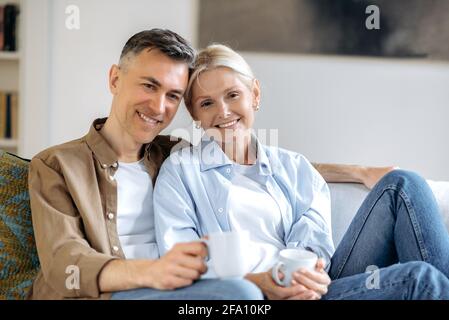 Ritratto felice carino anziano sposato caucasico coppia in abiti casual seduta su comodo divano in soggiorno, tenendo le tazze da tè, guardando la macchina fotografica, sorridendo. Marito e moglie riposano a casa Foto Stock