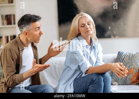 Incomprensioni, lite familiare. Sconvolto, indignato marito dai capelli grigi sta parlando emotivamente e guardando sua moglie, gesturando con le mani, moglie è sconvolto a causa di un dialogo, sperimentando stress Foto Stock