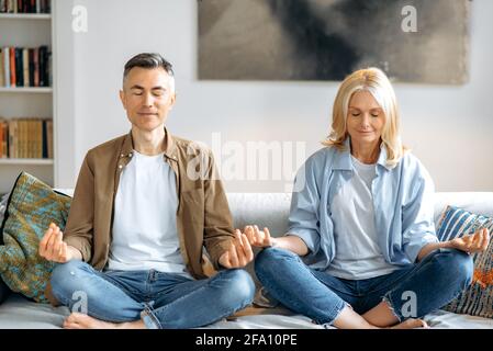 Sposato maturo coppia amorevole in abiti casual, che conduce uno stile di vita sano, facendo yoga a casa sul divano, seduto in posizione di loto in soggiorno, meditando, occhi chiusi, sorridente. Calma Foto Stock