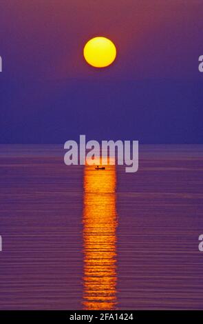 Il sole che sorge sul mare formando un lungo riflesso con una barca da pesca lontana nel centro. Puerto Pollensa, Maiorca, Spagna. Foto Stock