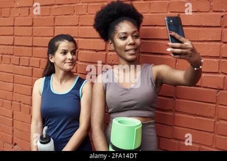 Immagine di donne atletiche giovani sorprendenti che prendono un selfie contro un muro di mattoni. Le donne nere sorridenti trascorrono il tempo insieme dopo l'allenamento sportivo. Foto Stock