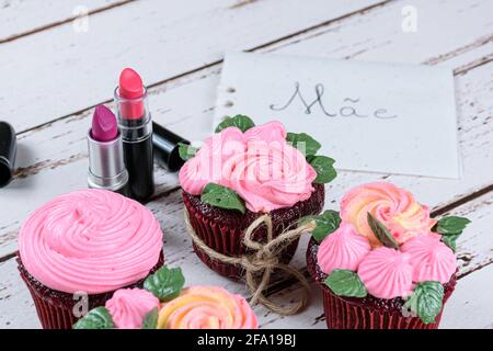 Cupcake di velluto rosso con glassa di crema al burro, accanto a due rossetti. In background nota scritta in portoghese, madre. Foto Stock