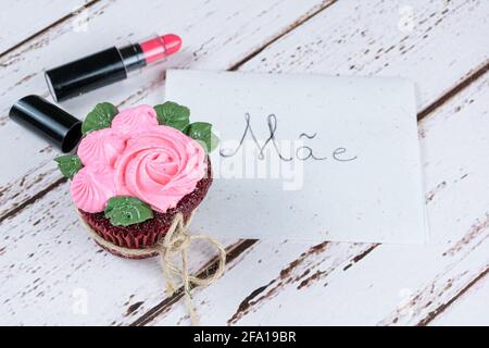 Cupcake di velluto rosso con glassa di crema al burro, accanto a un rossetto. In background nota scritta in portoghese, madre. Foto Stock