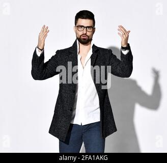 Giovane uomo con bearded elegante in occhiali e giacca tweed sta tenendo le mani in su e parla Foto Stock