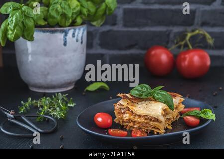 Pezzo di lasagna di soia vegetariana fatta in casa su un piatto nero, pomodori, erbe e basilico su sfondo scuro Foto Stock