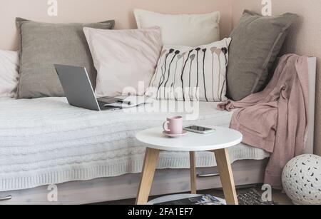 Camera accogliente con letto, cuscini grigi e rosa, tavolino da caffè, notebook, telefono cellulare e caffettiera preparati per una pausa, interni in stile scandinavo Foto Stock