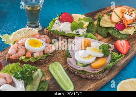 Varietà di panini danesi aperti su tavole di legno e sfondo blu, decorati con verdure, fragole e acquavit Foto Stock