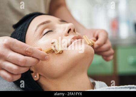 Cosmetician massaggiando il viso del cliente con massaggiatori a rullo in metallo ad acupressione Foto Stock