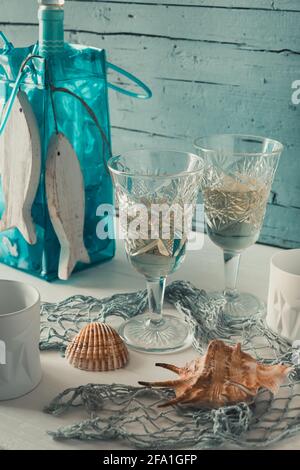Due bellissimi bicchieri ornamentali di vino bianco con decorazione marittima su un tavolo di legno bianco di fronte a sfondo di legno blu chiaro. Verticale. Foto Stock