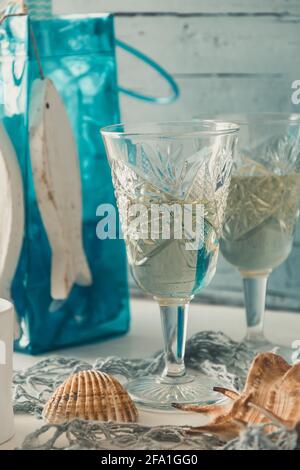 Due bellissimi bicchieri ornamentali di vino bianco con decorazione marina su un tavolo di legno bianco di fronte a sfondo di legno azzurro. Primo piano verticale Foto Stock