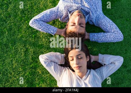 Vista dall'alto di una giovane coppia eterosessuale spensierata che riposa su un bel prato verde con le mani sotto la testa. Due persone che si rilassano godendosi insieme Foto Stock