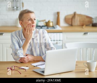 Donna matura frustrata che tocca la guancia, che soffre di mal di denti mentre si siede in cucina a casa mentre lavora da casa su un computer portatile, fuoco selettivo sulla donna. Concetto di salute dentale Foto Stock