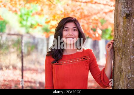 Sorridente ragazza biraciale in camicia rossa sorridente sotto il rosso acero in parco in autunno Foto Stock